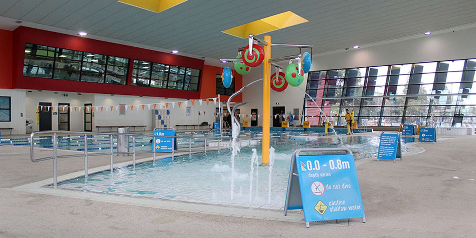 Indoor swimming pool featuring colorful decorations, shallow water areas, and safety signs, under a modern ceiling with large windows.