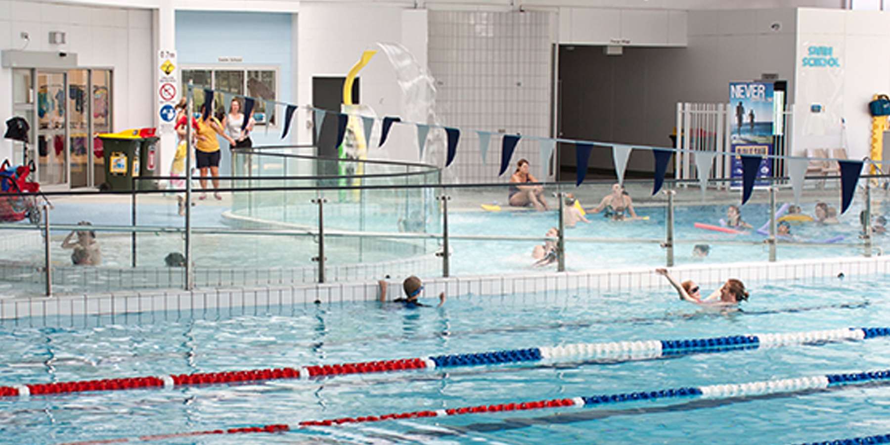 A busy indoor swimming pool, featuring children and adults swimming and playing, with colorful lane markers and a lifeguard on duty.