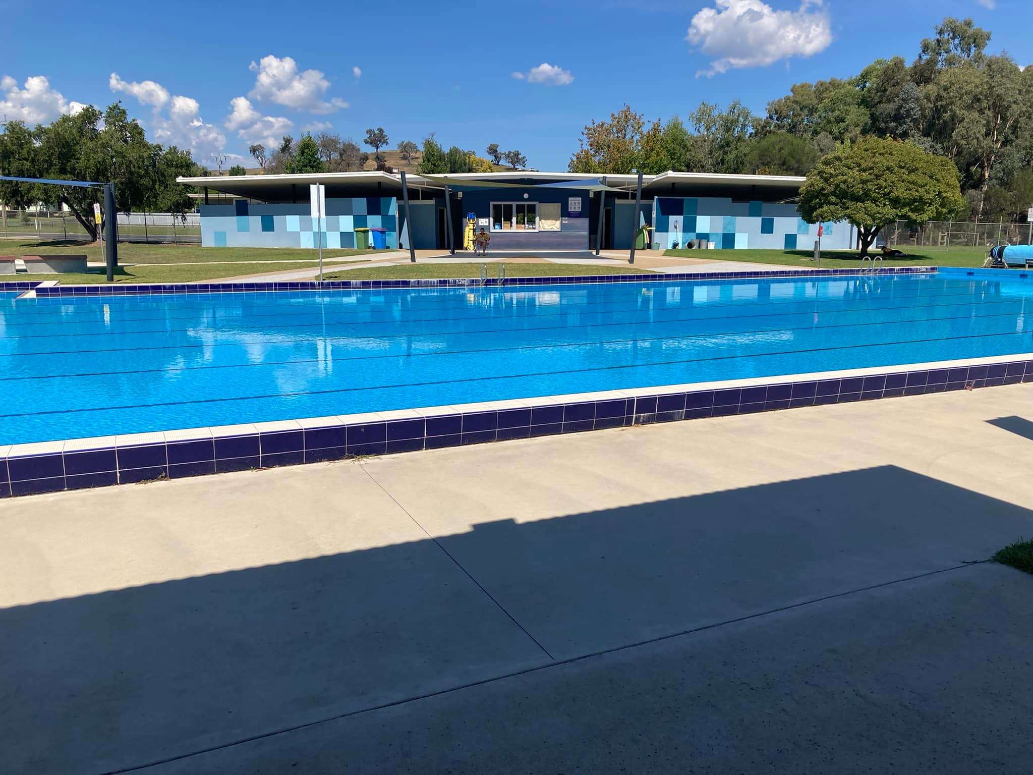 A large inground pool surrounded by green trees and grass and a blue brick changeroom in the background.