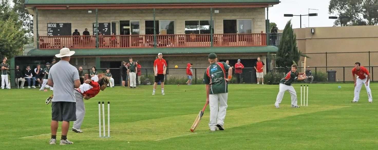 A team playing cricket with a building in the background and one person bowling the ball