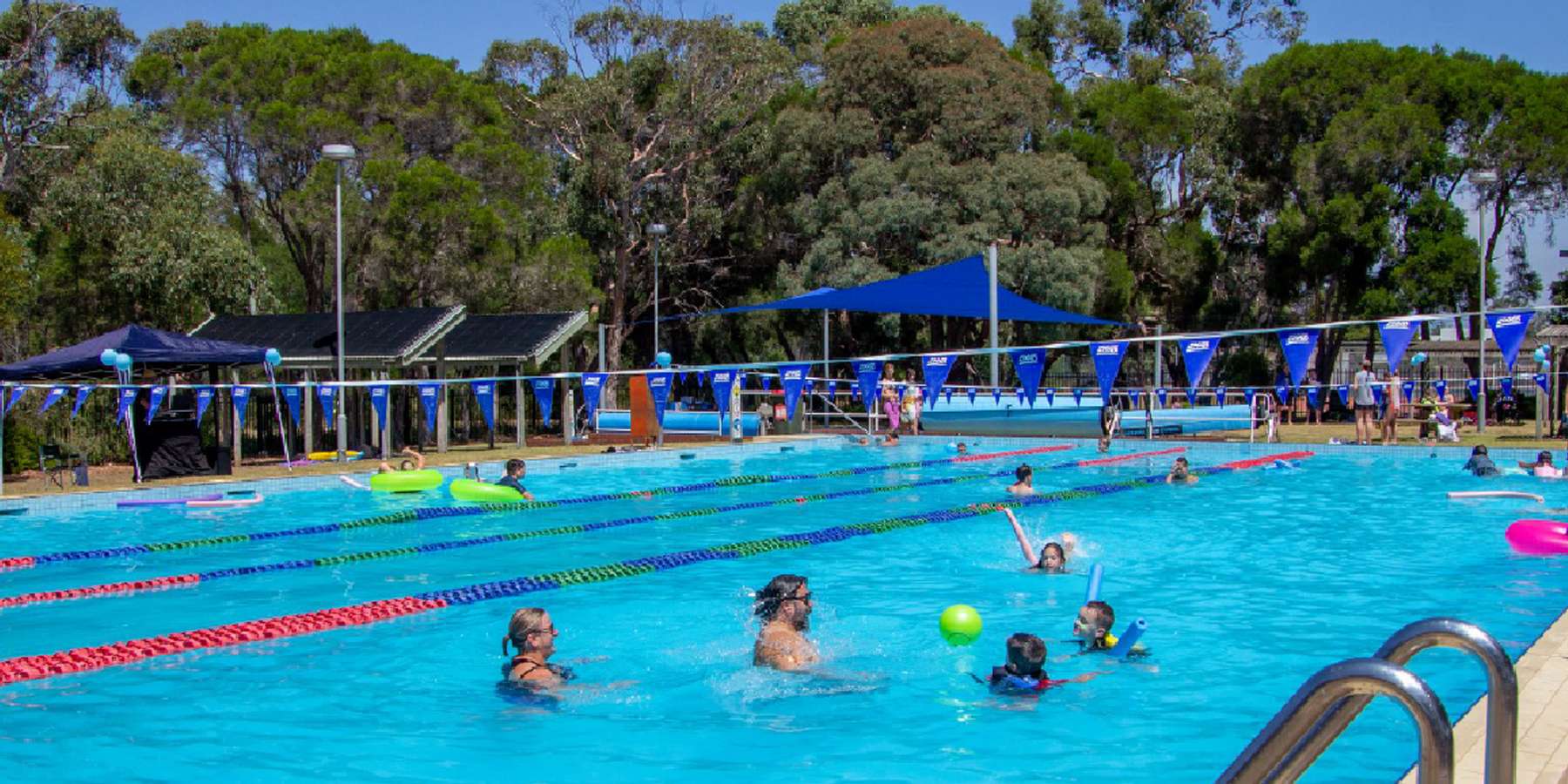 Children swim and play in a bright blue pool surrounded by trees and shaded areas, enjoying a sunny day.