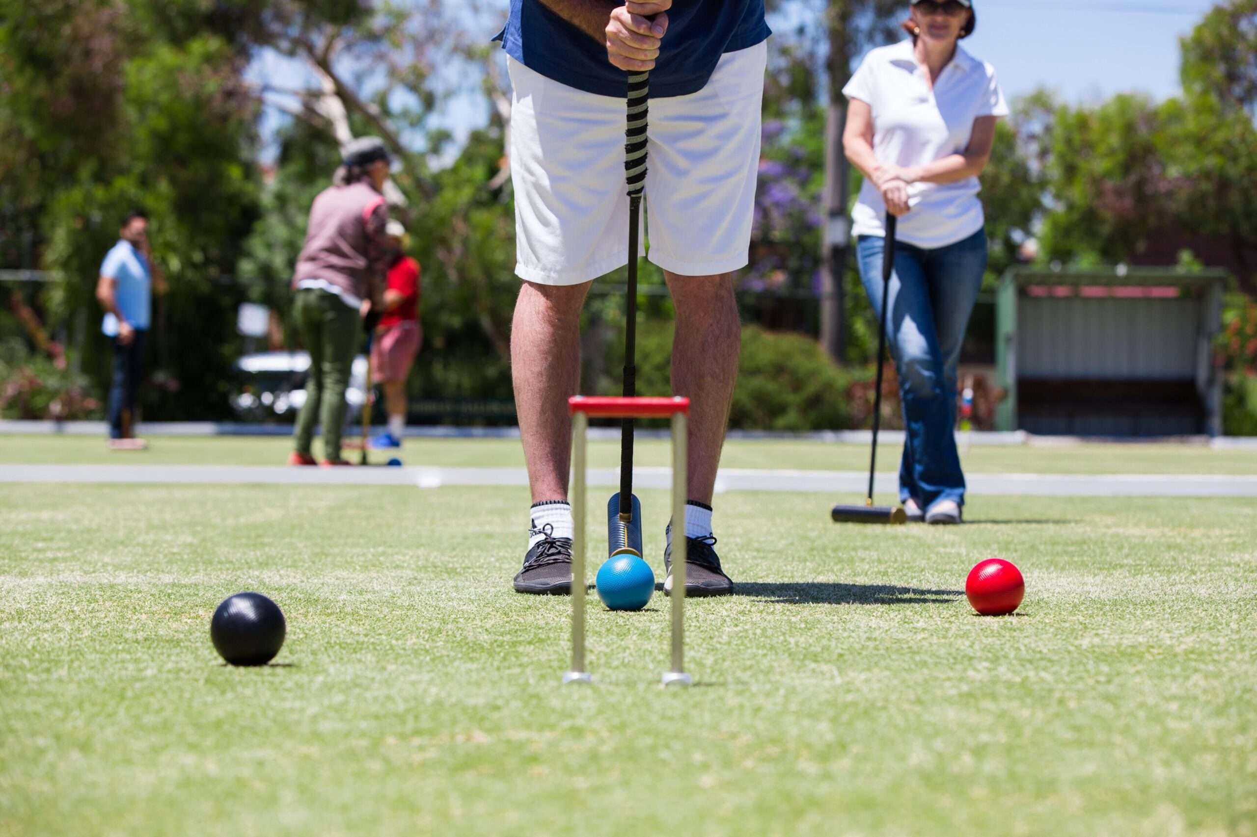 A person stands on a vibrant green croquet lawn, ready to hit a blue ball with a mallet. The player is wearing white shorts and a blue shirt, with the mallet poised above the ball. Nearby, a red ball and a black ball are visible on the grass. In the background, other players are engaged in their own games, enjoying a sunny day.