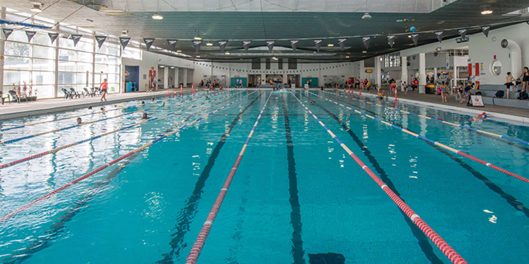 Indoor swimming pool with clear blue water, multiple lanes, and swimmers engaged in various activities along the sides.