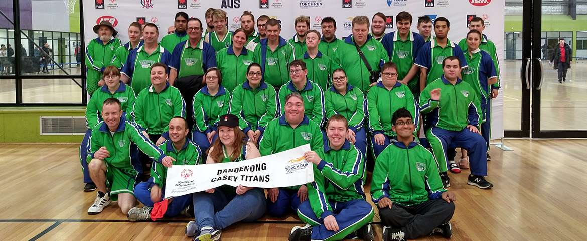 A large group of athletes wearing green jackets poses together, holding a banner in a sports venue decorated with sponsor logos.