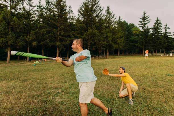 A man swings a green bat while a woman in a yellow shirt crouches, ready to catch a ball in a grassy field surrounded by trees.