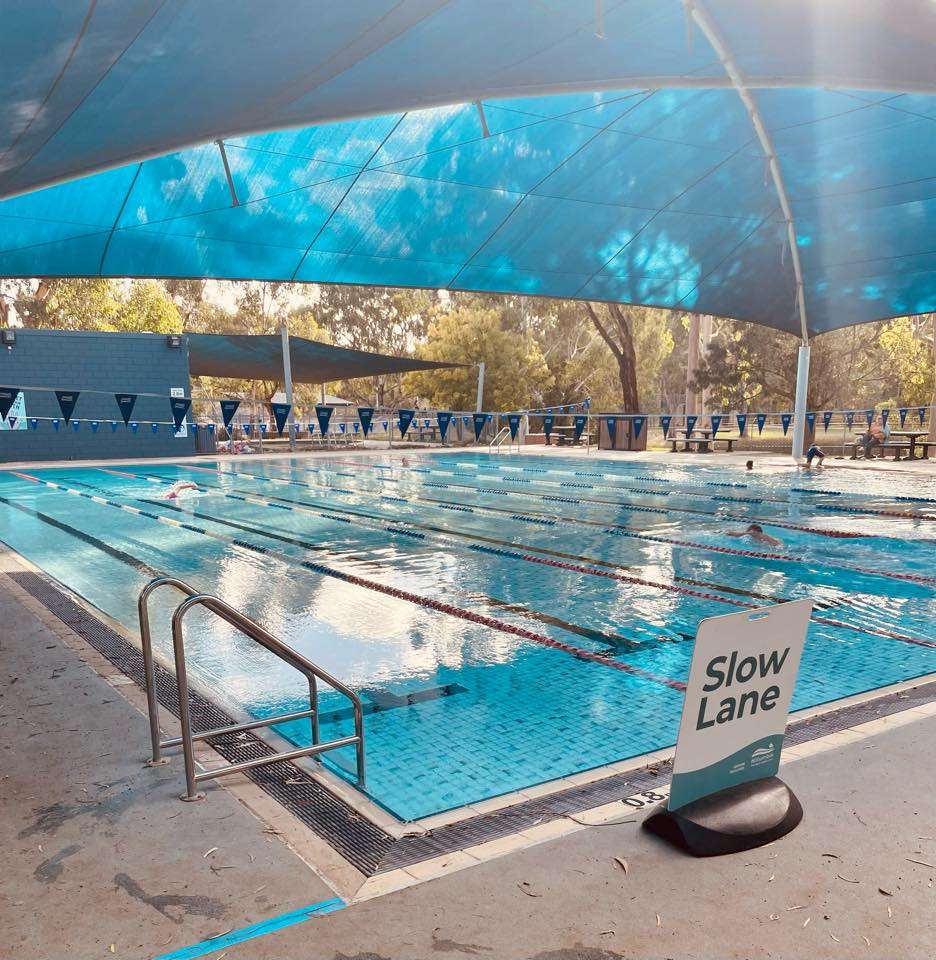 A large inground pool surrounded by green trees and grass and a blue brick changeroom in the background with a large blue shade sail.