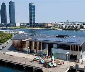 Modern waterfront building with solar panels, surrounded by boats, and skyscrapers in the background under a clear blue sky.