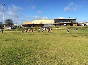 A grassy field with children playing soccer, a building in the background, and clear blue skies.