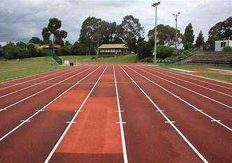 An empty athletic track with red lanes, surrounded by green trees and a pavilion in the background, under a cloudy sky.