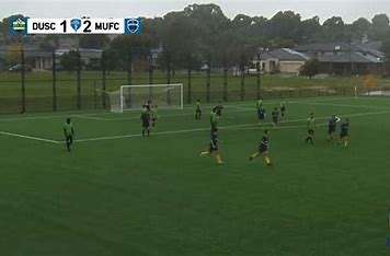 Players in green and blue jerseys compete on a wet soccer field, with a score of DUSC 1, MUFC 2 displayed above.