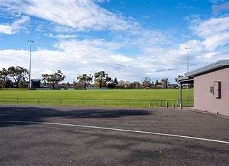 A sunny sports field with green grass, surrounded by trees and under a blue sky, adjacent to a gray building.