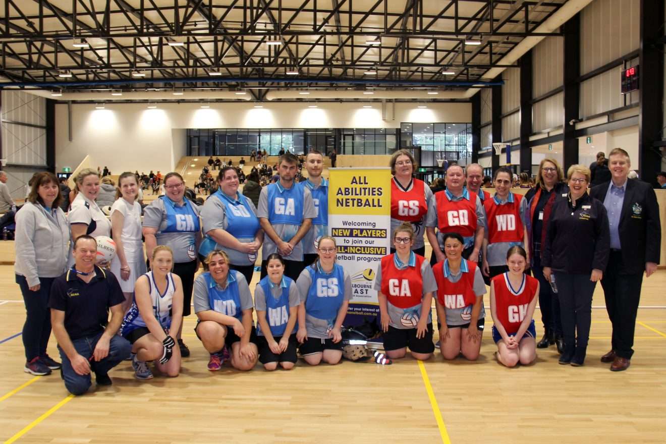 Photo of a group of men and women posing for the camera. On the right side there are players in blue netball bibs, and the on the left side there are players with red bibs