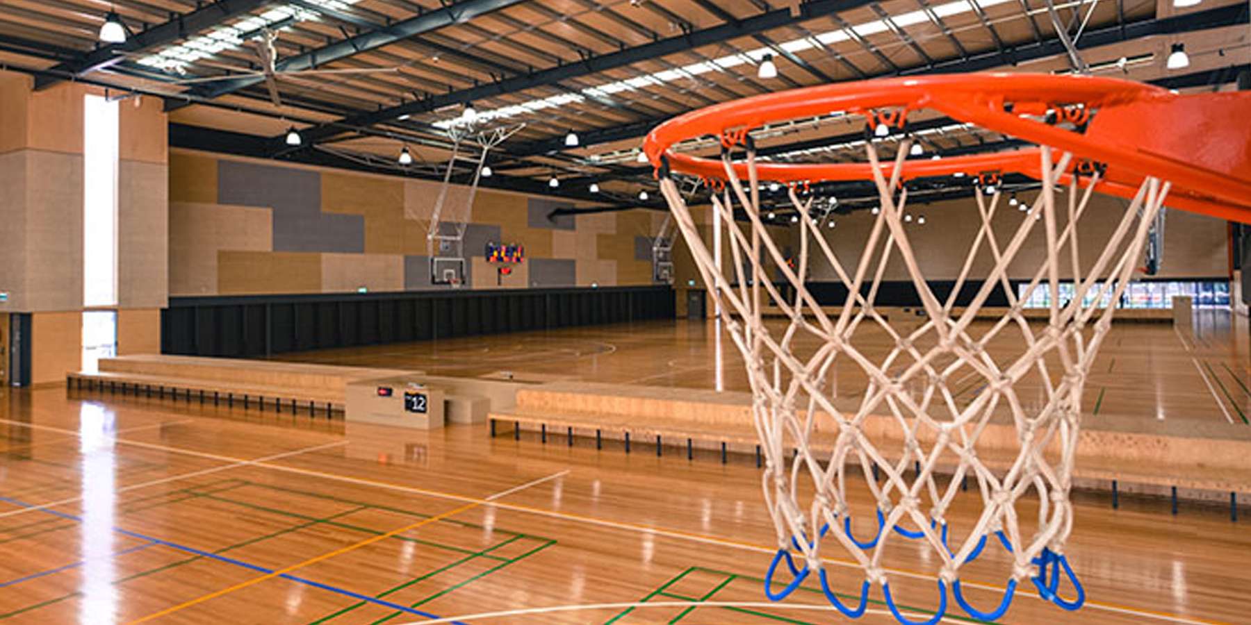 A close-up view of a basketball hoop in a spacious indoor gymnasium with polished wooden floors and bright overhead lights.