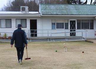 Image displays a green flat croquet club, there is a player with their back to the camera holding a croquet club, playing a game. There is a building in the background.