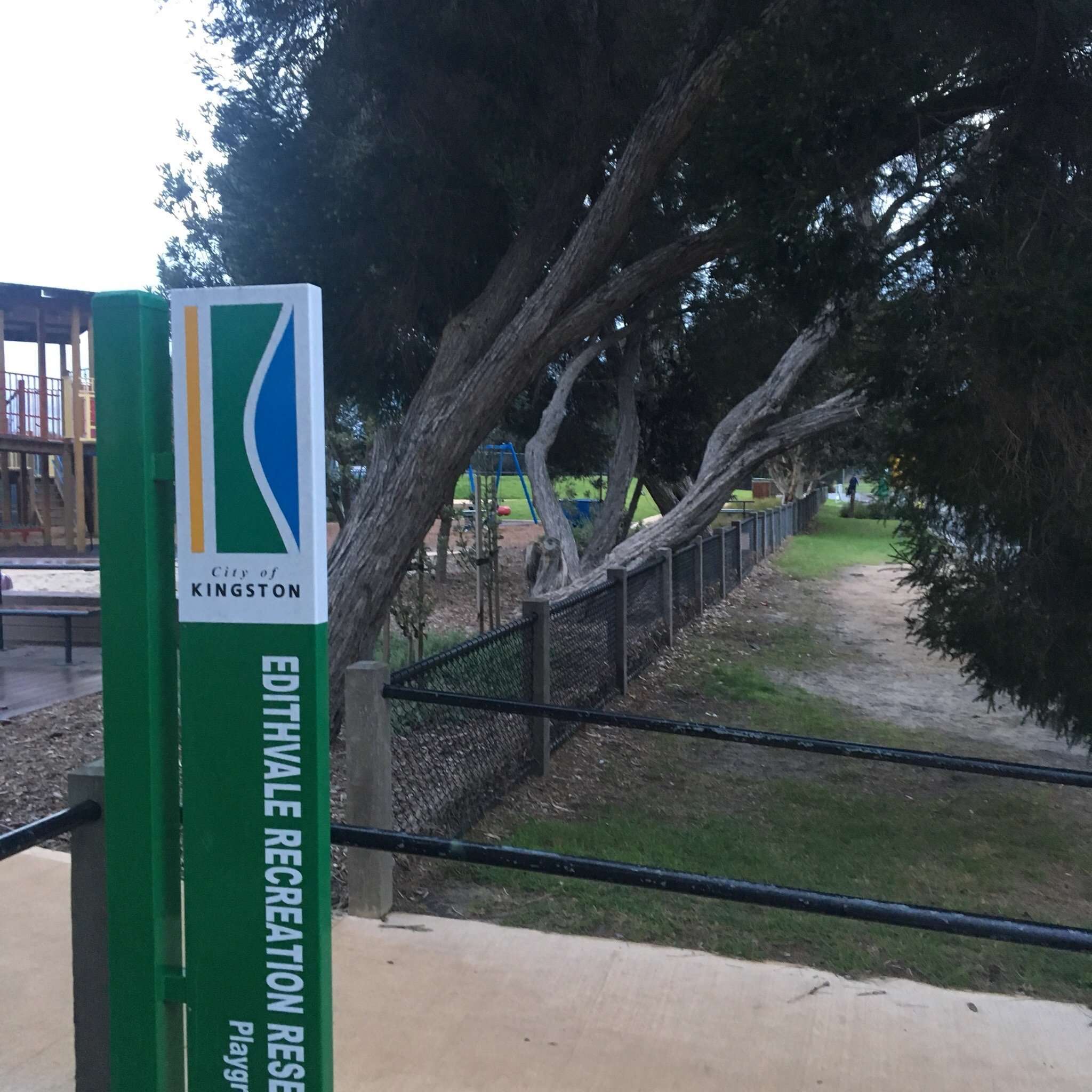 Photo displays a sign that reads Edithvale Recreation and in the background you can see overhanging trees and a fence, with a path leading into the recreation reserve
