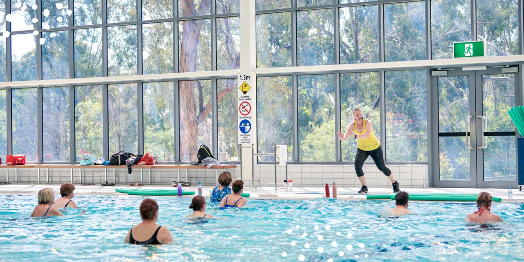 An instructor leads a water aerobics class in a bright indoor pool, surrounded by participants in the water.