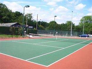 A bright tennis court with green and red surfaces, surrounded by trees and a building in the background under a partly cloudy sky.