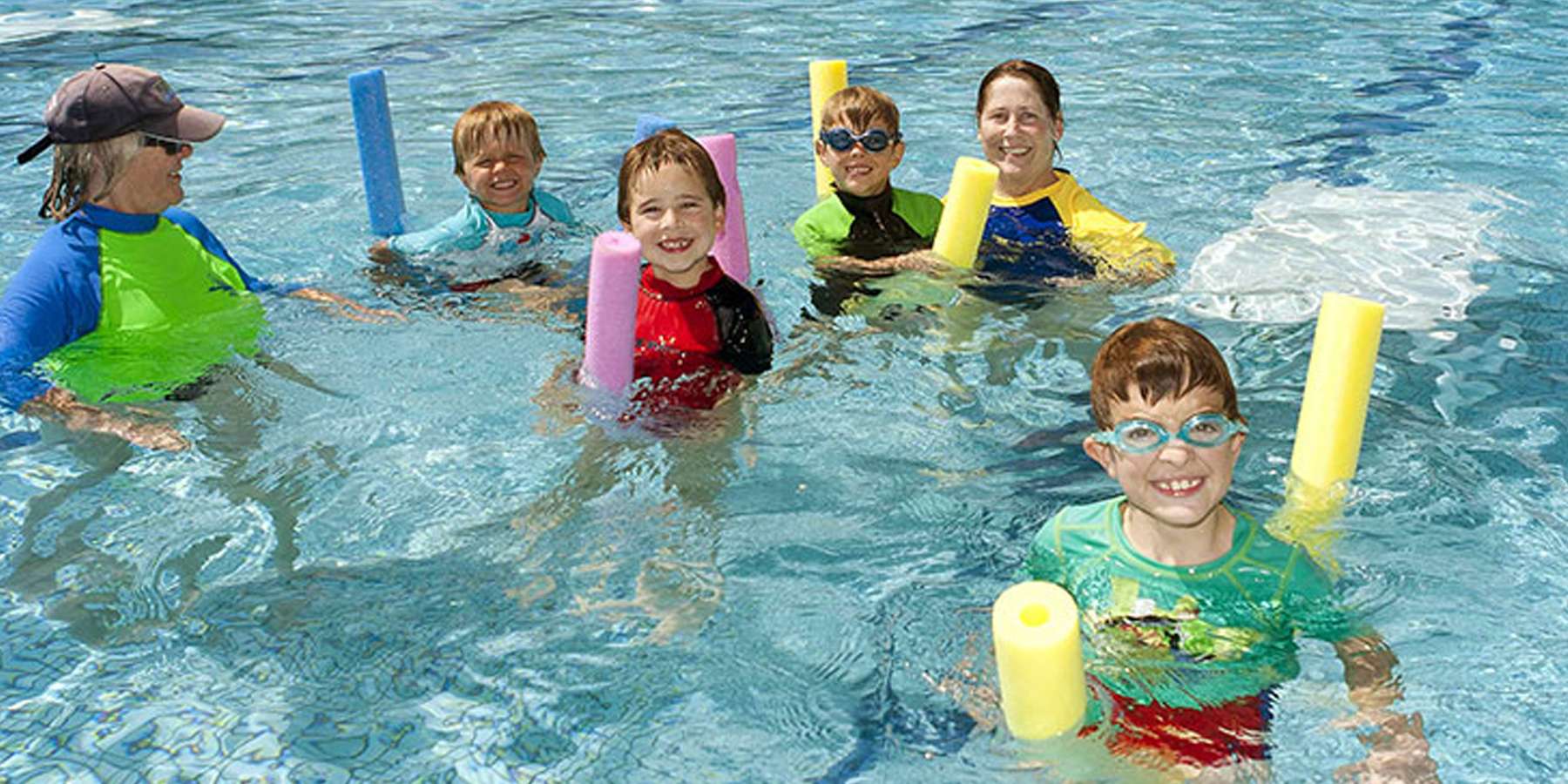 A group of children and an instructor enjoy swimming in a pool, using colorful pool noodles for support and safety.