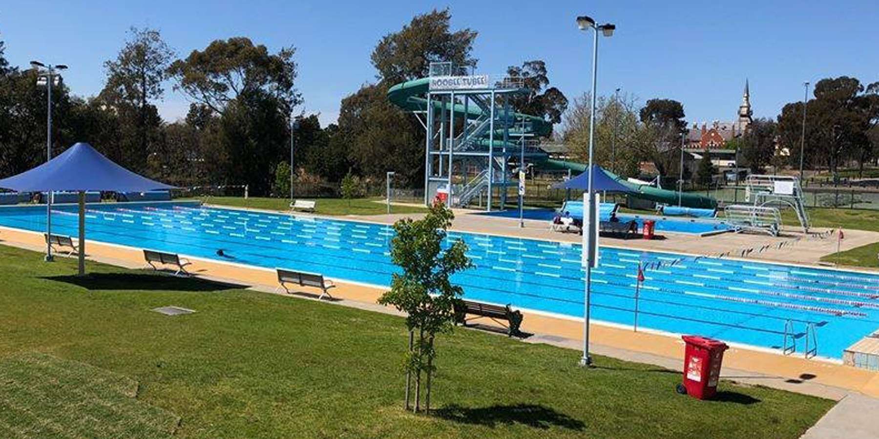 A sunny outdoor swimming pool with lanes, diving boards, shaded areas, and landscaped greenery surrounding it.