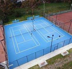 Image displays multiple blue turf tennis courts from above. Courts have white lines marking them, and black fencing around the outside, there is parkland and trees behind the courts.