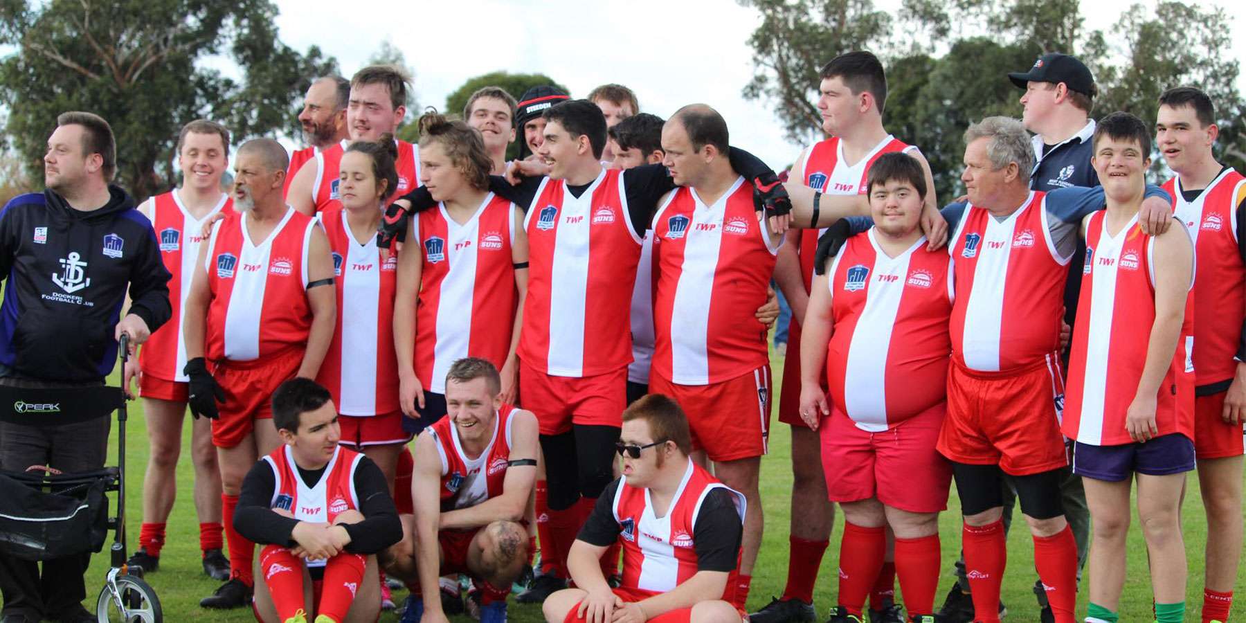 A large group of athletes in red and white sports uniforms smiles together on a green field, celebrating team spirit and camaraderie.
