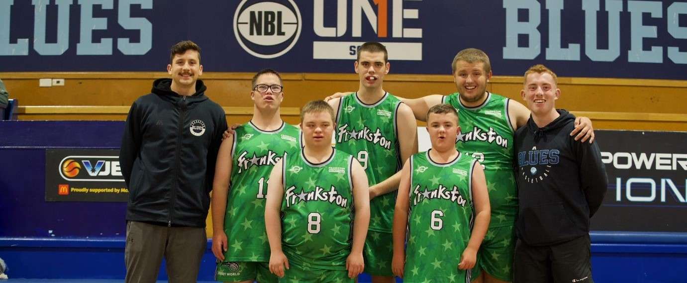 A basketball team poses together, wearing green jerseys, with two coaches in front of a basketball court backdrop.