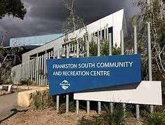 Image displays the Frankston South Community Centre in the background, there is a blue sign out the front, there are a few trees around the building and small shrubs in front of the blue sign