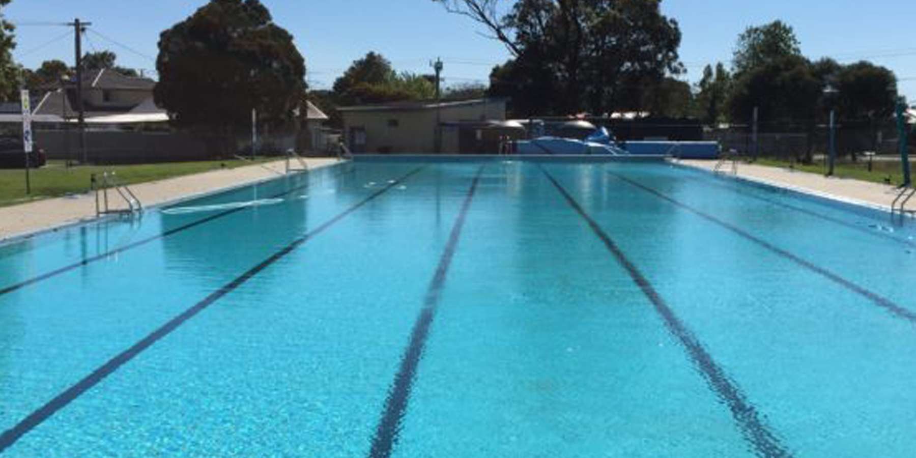 A bright outdoor swimming pool with clear blue water and lane markers, surrounded by trees and a clear blue sky.