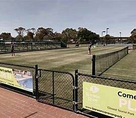 Image displays tennis multiple tennis courts with astro turf surfaces. Small black fencing around the courts, and you can see people in a distance playing tennis. The sky is blue and there are trees way off in the distance.