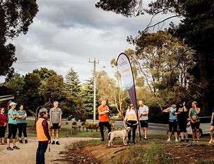A group of runners, some with a dog, gather near a branded flag in a park under cloudy skies, preparing for an event.