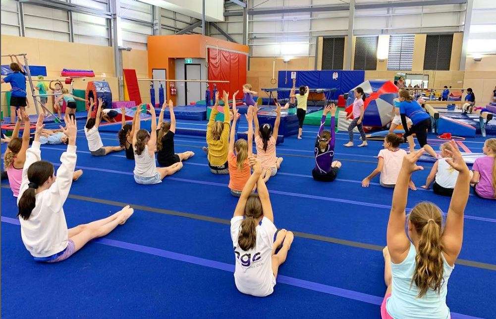 A group of young gymnasts stretches on blue mats in a gymnasium, with colorful equipment in the background.