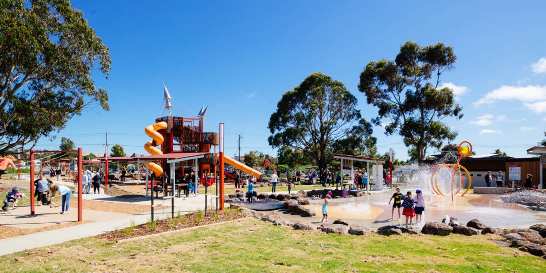 A vibrant public playground featuring a slide, climbing structures, and a splash pad with children playing under a clear blue sky.