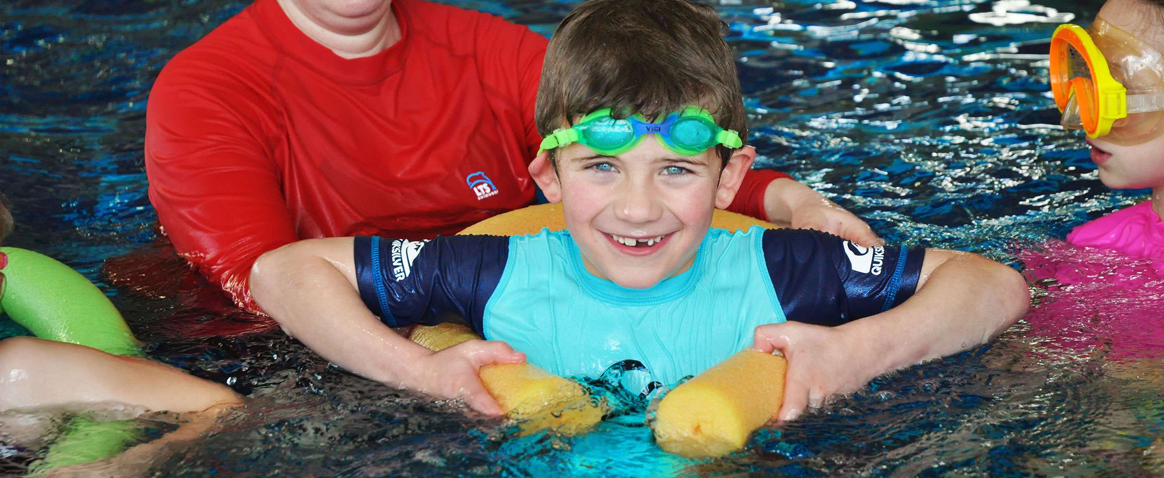 A young boy with swimming goggles and blue swimming top in the pool with instructor wearing a red top.