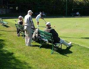 Image displays a green flat croquet club, there are players with their back to the camera holding a croquet club, playing a game, and a few people sitting on green benches.
