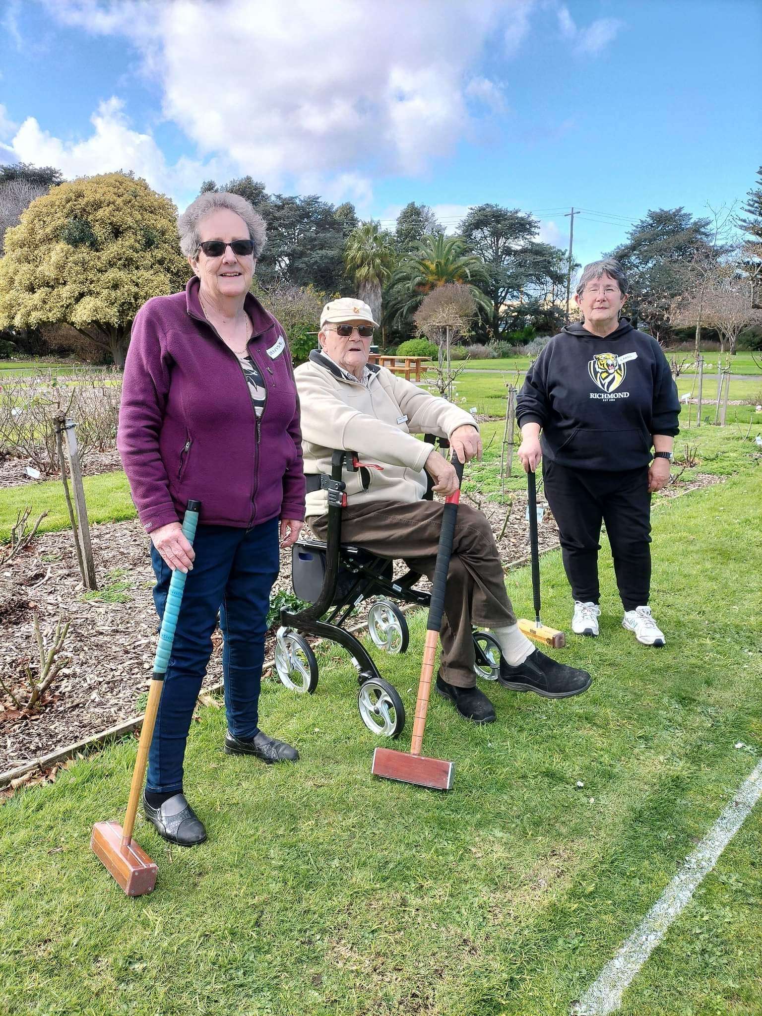 Three people stand in a garden, holding mallets, with a wheelchair nearby, under a cloudy sky and surrounded by greenery.