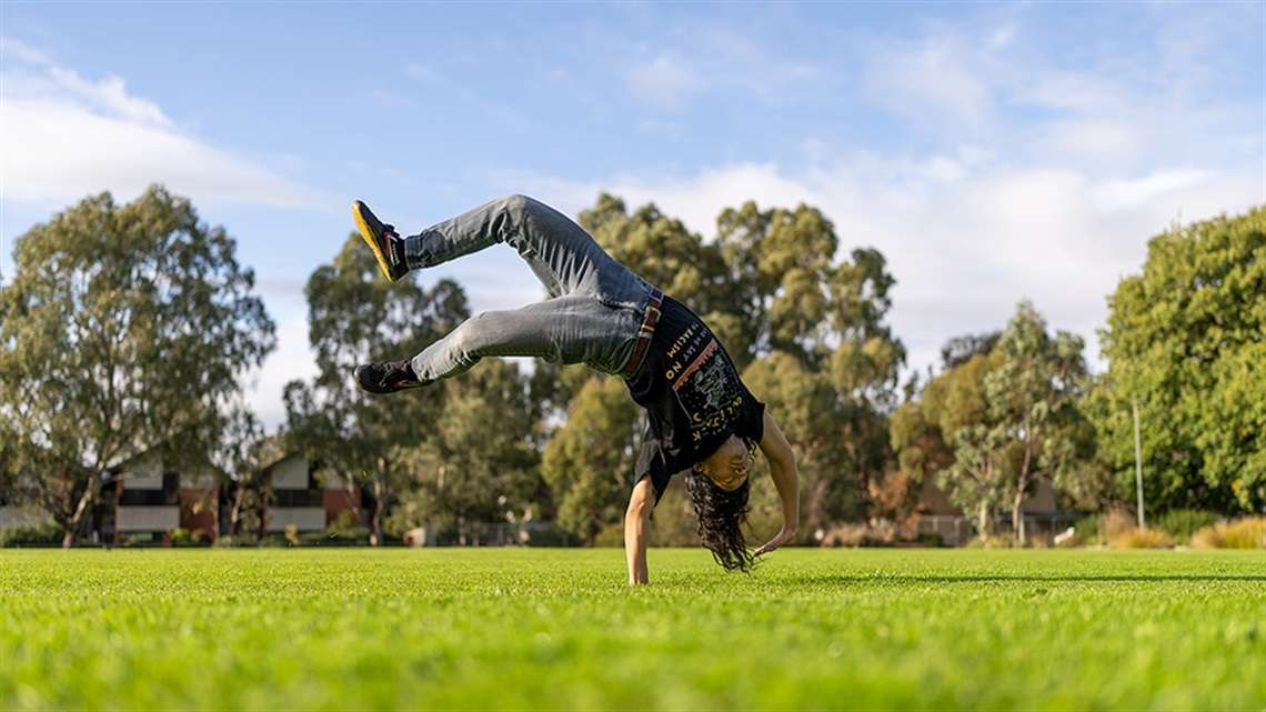 a large park with green grass and surrounded by trees with a boy doing cart wheels.