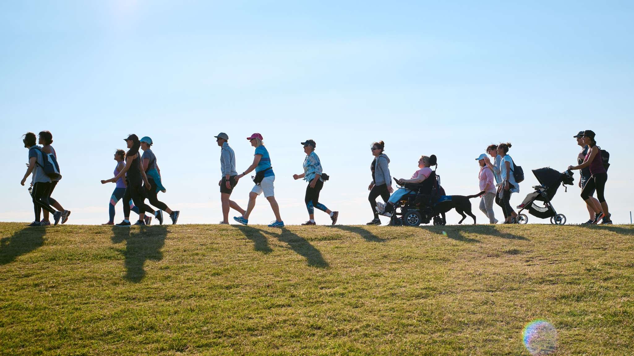 15 people walking outdoors in front of blue sky. Includes various ages, wheelchair, guide dog, and pram.