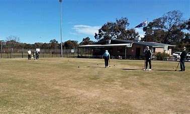 Image displays a green flat croquet club, there are players with their back to the camera holding a croquet club, playing a game. There is a building in the background.