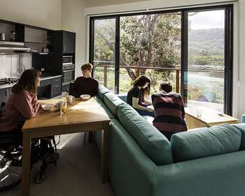 Image displays four people sitting accommodation with a nice view. Two people are sitting on a blue couch and two people are sitting at a dining room table