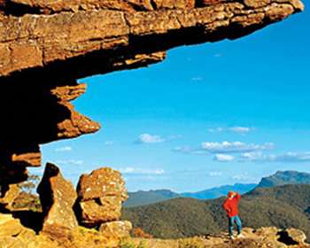 Image of a huge rock face with a view out to a blue sky with white clouds, and hills covered in trees in the distance. There is one person in the distance in a red jacket and blue pants.
