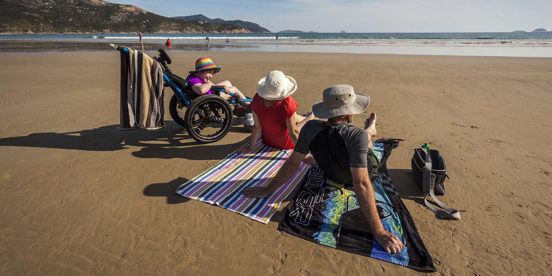 Image displays one person in a beach wheelchair and two people sitting on towels on the sand looking out towards the ocean