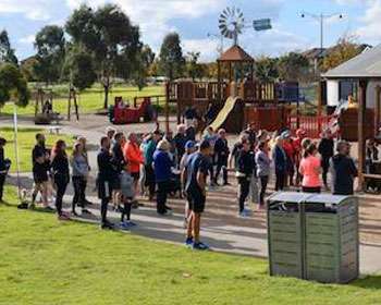 Group of people standing facing a pergola and someone presenting. There is a playground in the background