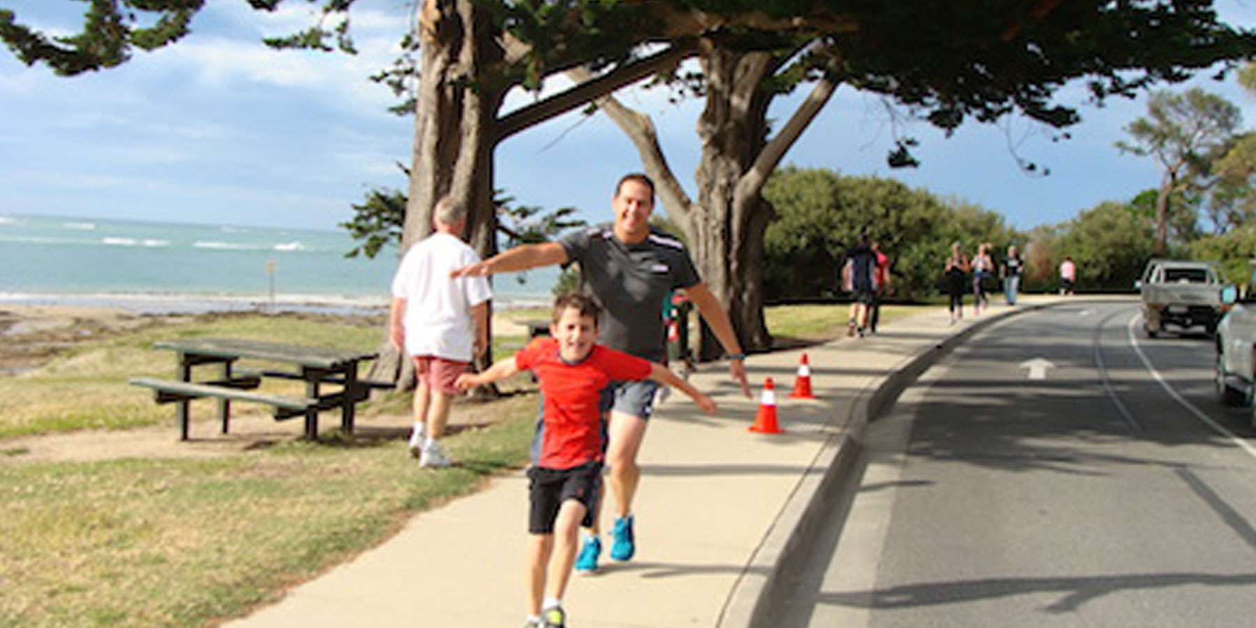 Image of and man and child running along a footpath with their arms outstreached. the beach is in the background