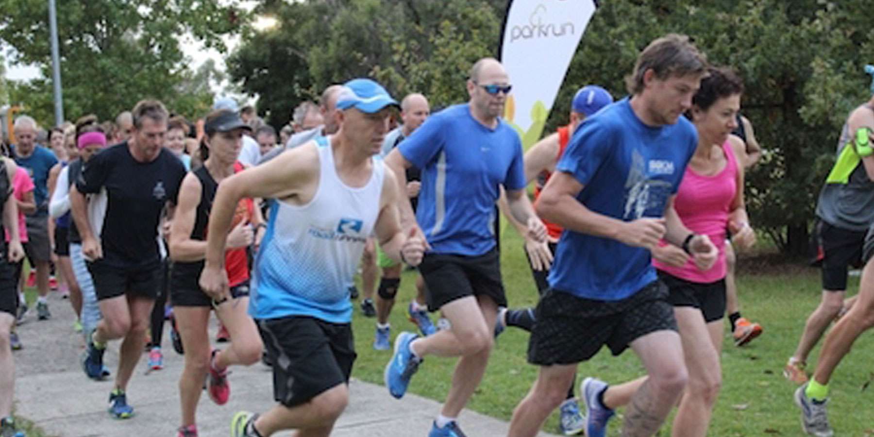 A diverse group of runners in athletic gear starting a race on a path, surrounded by greenery and spectators.