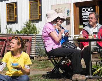 Image of a woman with a dog sitting on the ground holding a cup. A group of three people are sitting at a table in front of a shop in the background
