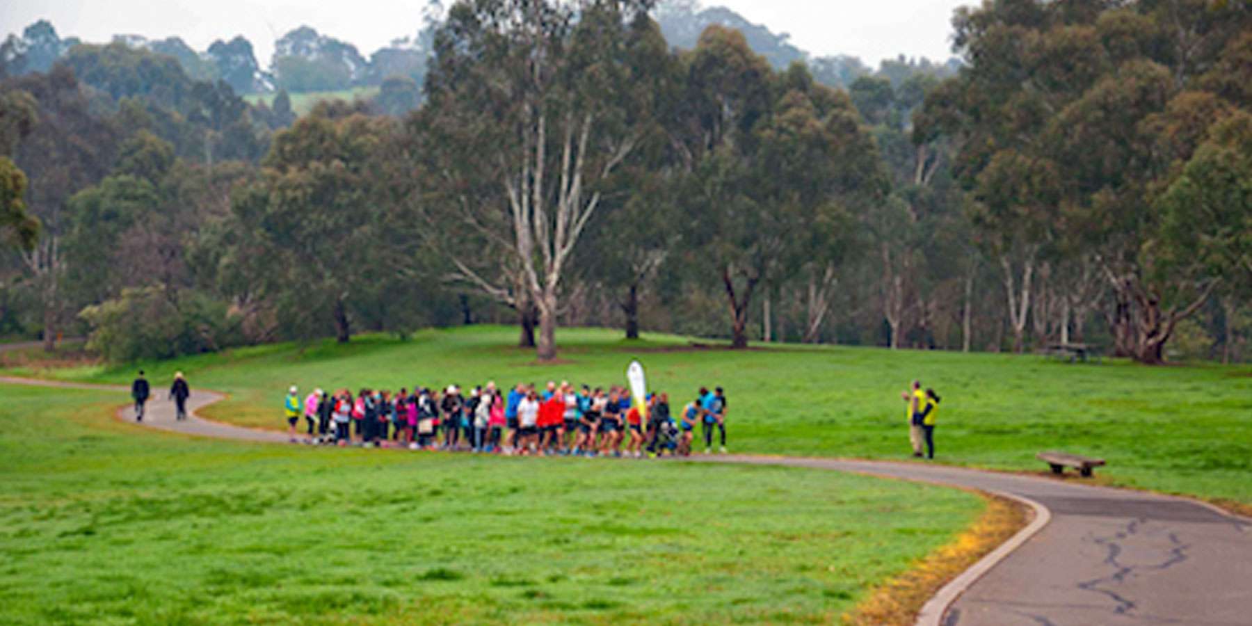 Green open space with trees in the background and a group of people running along a path
