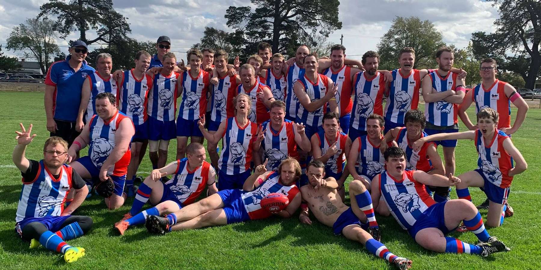 A jubilant team of male football players in red, white, and blue uniforms poses together on a sunny field, celebrating their victory