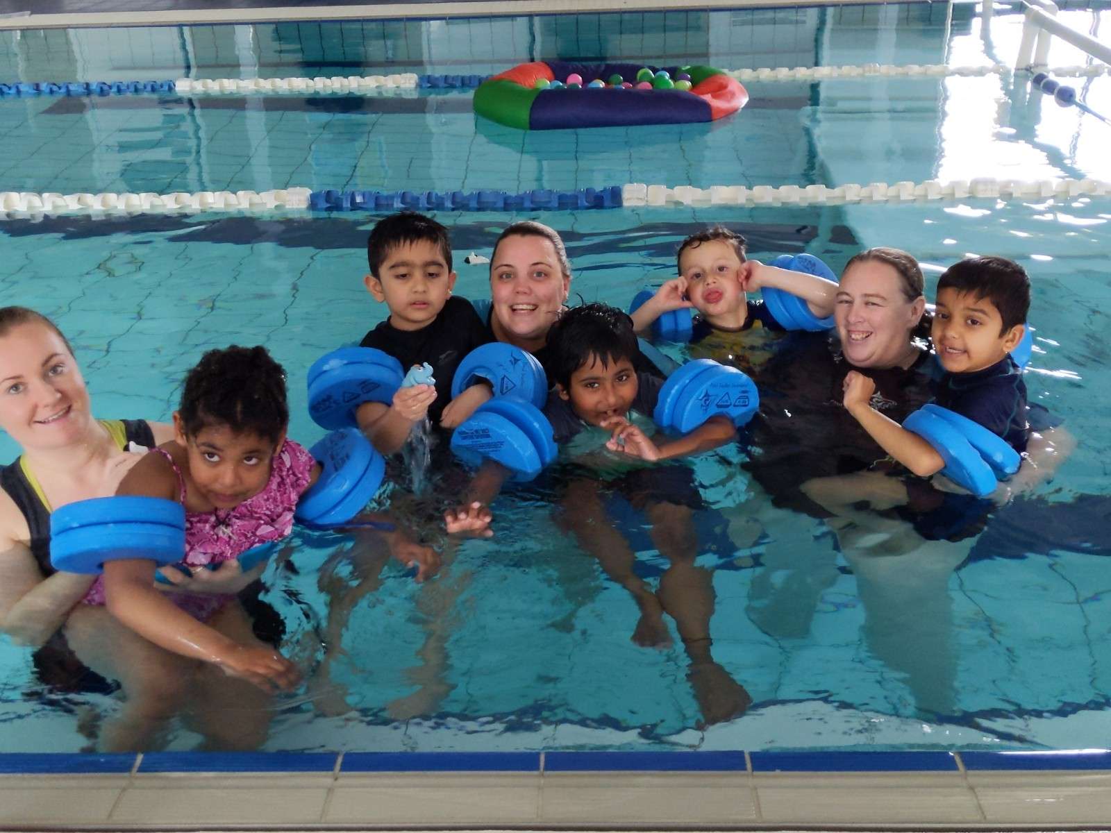 A group of seven children and two adults smile together in a swimming pool, wearing brightly colored life jackets.