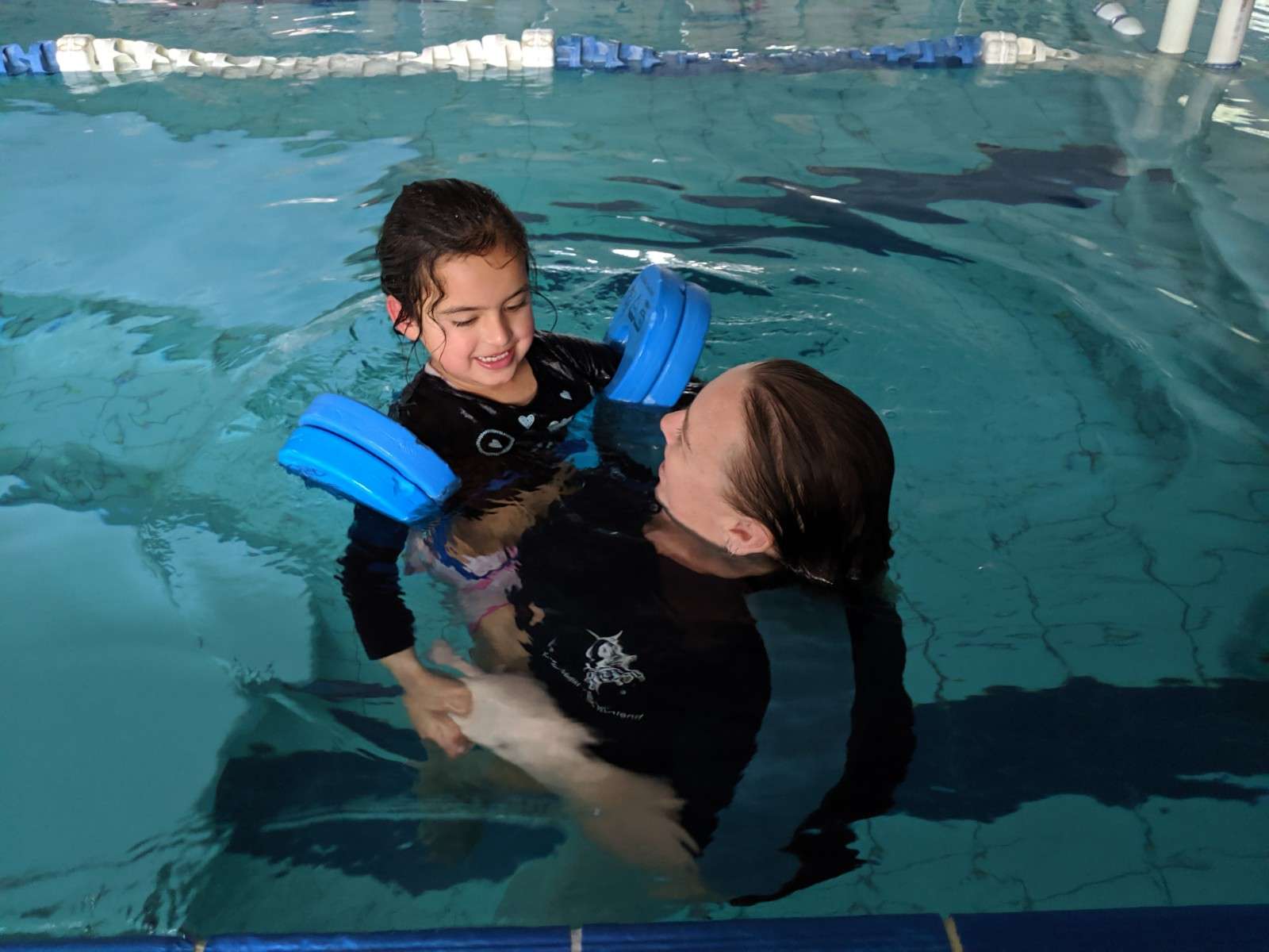 A swimming instructor supports a child in a pool, with the child wearing floatation wings for safety. The water is calm and inviting.