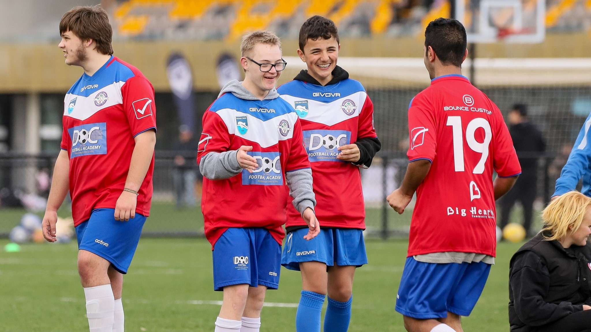 Four young soccer players in red and blue jerseys converse on the field, with soccer balls and spectators in the background.